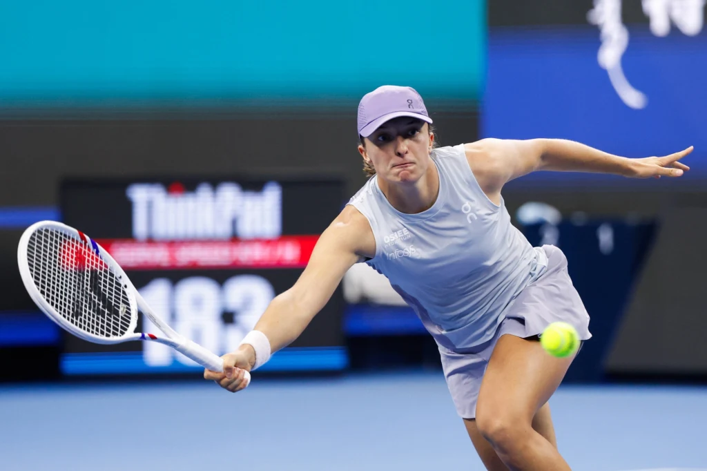 Tenisistka w jasnym stroju sportowym wykonuje zamach rakietą w trakcie dynamicznego meczu na twardym korcie, skupienie widoczne na twarzy zawodniczki i żółta piłka tuż przed jej rakietą.