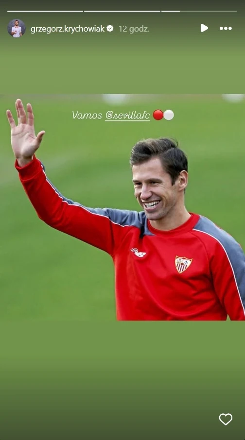 Uśmiechnięty mężczyzna w czerwonym dresie z herbem klubu Sevilla FC, stojący na murawie boiska i machający ręką w geście pozdrowienia.