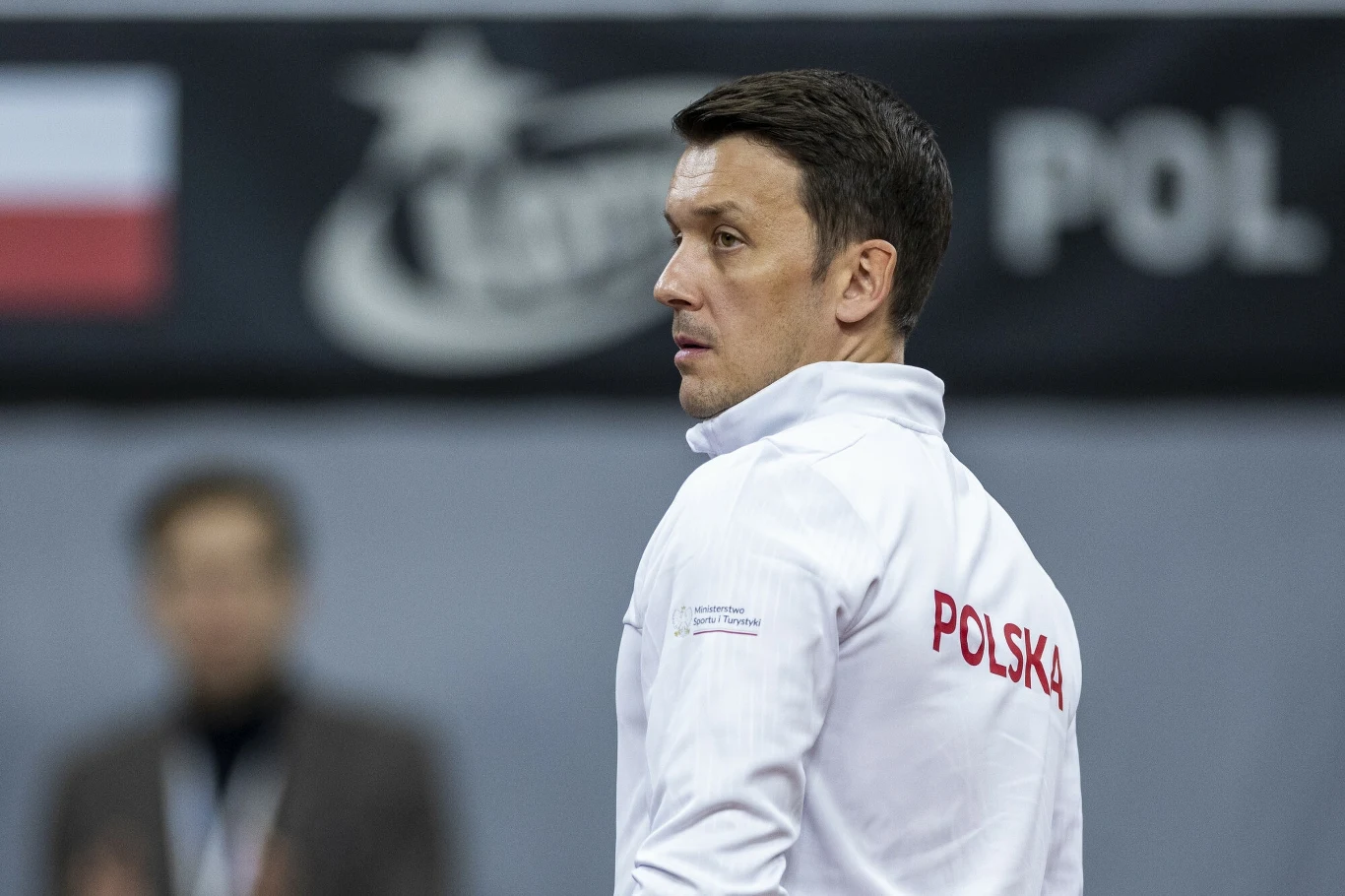 Mężczyzna w białej sportowej kurtce z napisem 'Polska' na plecach, stojący bokiem na tle rozmytej flagi Polski oraz napisu 'POL', sugerujących oficjalną reprezentację kraju.