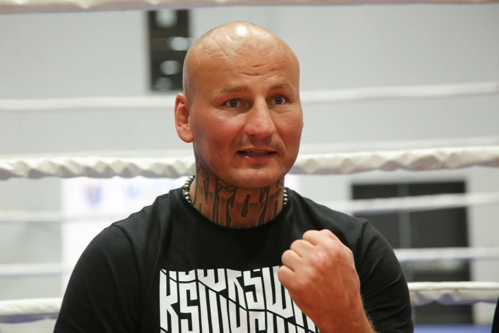 Mężczyzna w czarnej koszulce z widocznym tatuażem na szyi stoi na tle lin bokserskiego ringu, zaciśnięta pięść uniesiona w charakterystycznym geście.