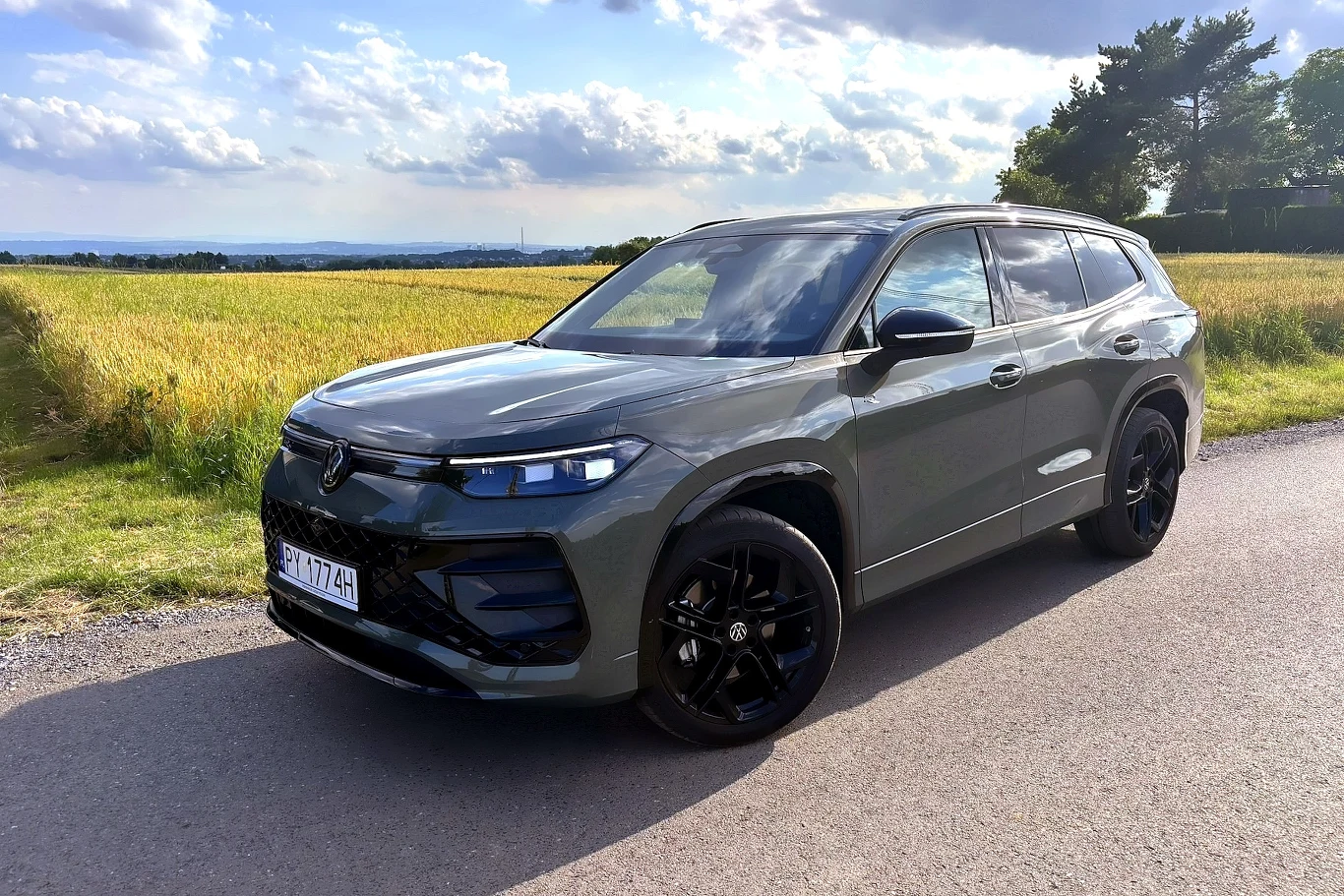 Volkswagen Tayron to nowy model, obecny na rynku dopiero od roku / Fot: Tomasz Szpil Nowoczesny SUV w zielonym kolorze stoi zaparkowany na wiejskiej drodze, otoczony polami zboża i delikatnie zachmurzonym niebem, jego czarne felgi wyróżniają się na tle asfaltu.