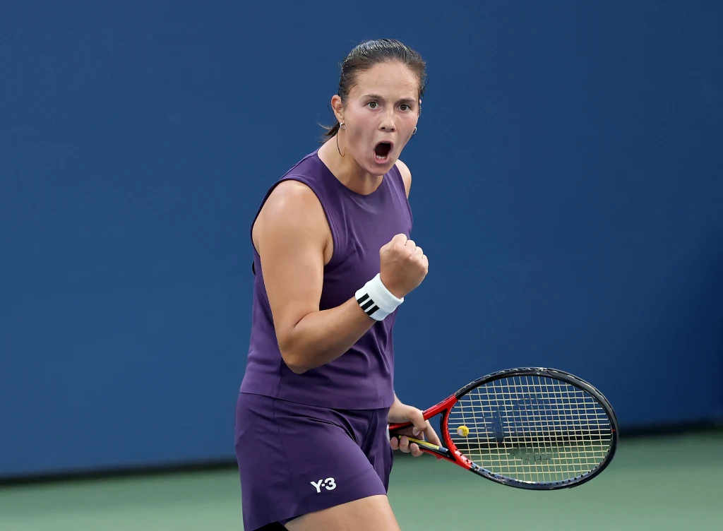 Tenisistka w sportowym stroju stoi na korcie z zaciśniętą pięścią i wyrazem determinacji na twarzy, trzymając rakietę tenisową w lewej ręce i wyrażając emocje zwycięstwa.