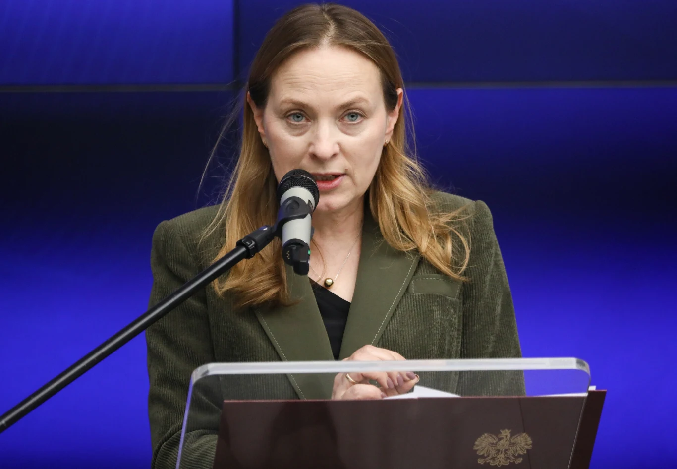 Minister funduszy i polityki regionalnej Katarzyna Pełczyńska-Nałęcz Kobieta w zielonej marynarce przemawia do mikrofonu, stojąc za przezroczystą mównicą z godłem państwowym, na tle niebieskiego ekranu.