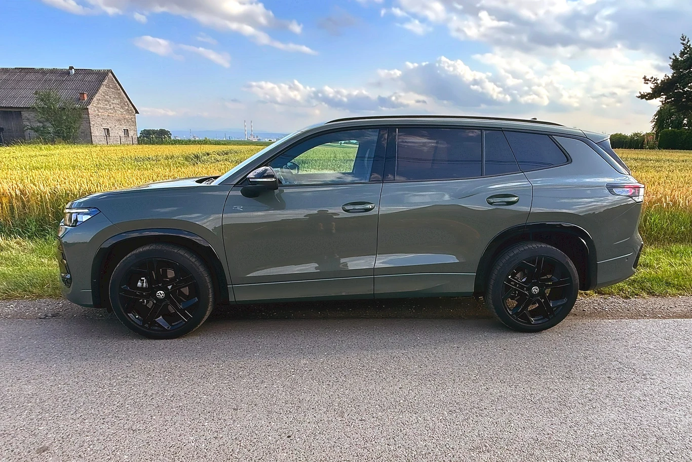 Tayron ma spory rozstaw osi, o 11 cm większy niż Tiguan / Fot: Tomasz Szpil Szary SUV zaparkowany na poboczu asfaltowej drogi, w tle pole uprawne oraz budynek gospodarczy na skraju wiejskiego krajobrazu, na niebie widoczne chmury.
