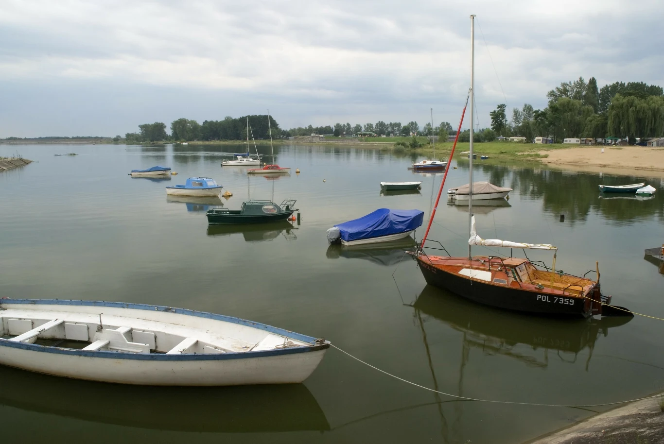 Jezioro Otmuchowskie Kilka małych łodzi i żaglówek cumuje na spokojnej wodzie przy brzegu, wokół widać zielone drzewa oraz w oddali piaszczystą plażę, a niebo jest lekko zachmurzone.