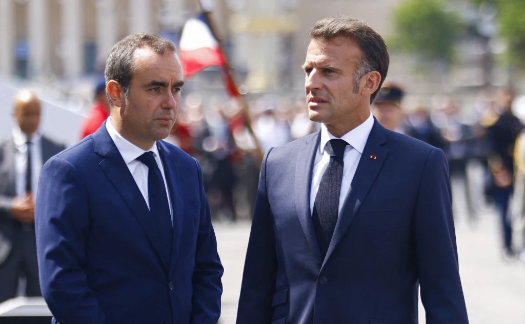 Premier i prezydent Francji: Sebastien Lecornu i Emmanuel Macron Dwóch mężczyzn w garniturach rozmawia ze sobą na zewnątrz podczas oficjalnego wydarzenia, w tle widoczne postaci i rozmazana flaga w barwach narodowych.