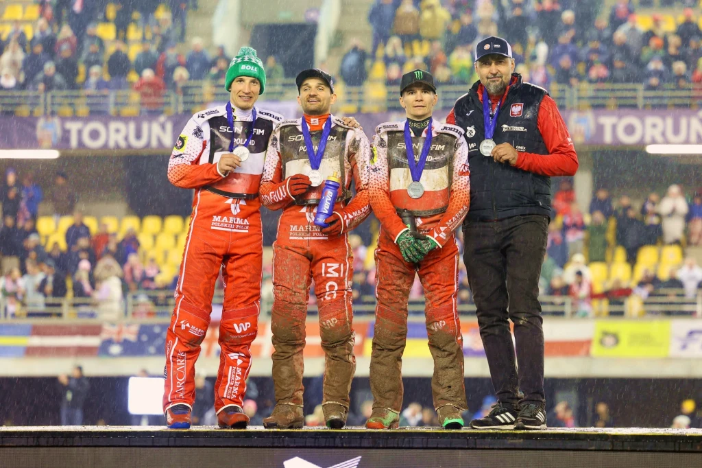 Kadra Polski na podium SoN w Toruniu Trzech sportowców w kombinezonach i kaskach żużlowych pozuje do zdjęcia na podium z medalami na szyjach, obok nich stoi trener ubrany w kurtkę i czapkę, a w tle znajdują się kibice na trybunach stadionu.