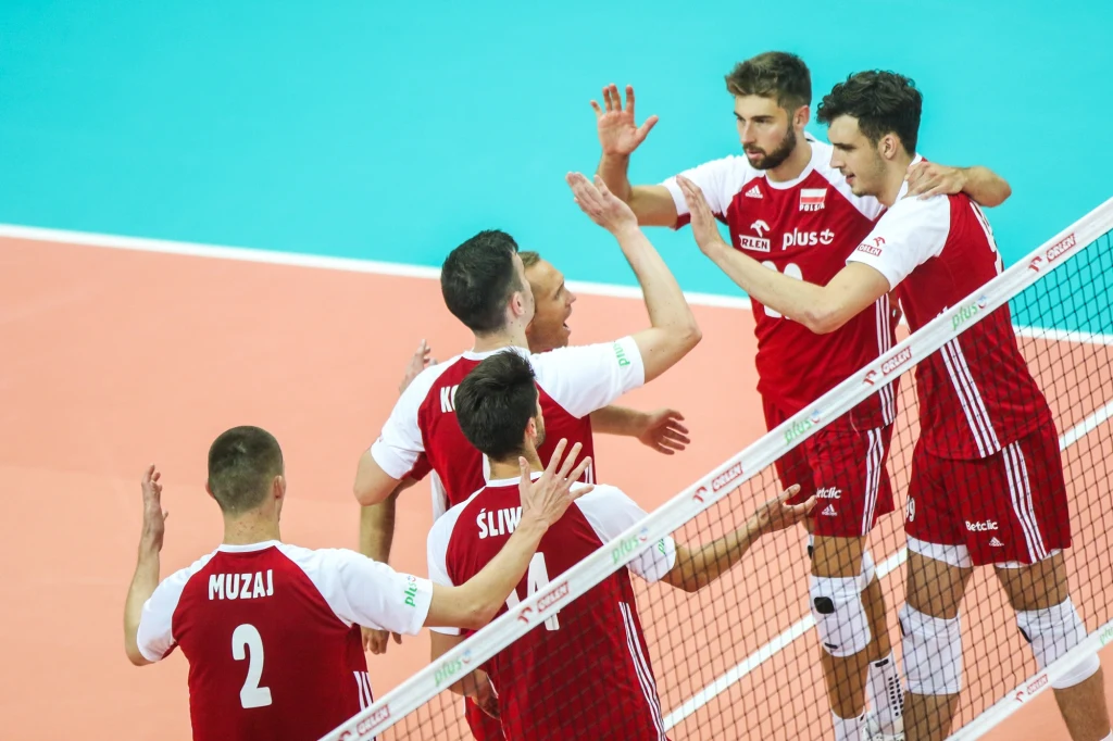Maciej Muzaj w reprezentacji Polski Grupa siatkarzy ubranych w czerwono-białe stroje świętuje zdobycie punktu na boisku, przybijając sobie piątki obok siatki, sportowa atmosfera podkreślona jasnym oświetleniem i kontrastującą niebieską podłogą.