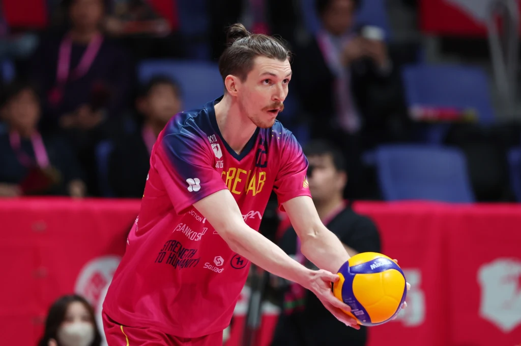 Maciej Muzaj Zawodnik drużyny siatkarskiej w różowo-niebieskim stroju szykuje się do wykonania zagrywki, trzymając żółto-niebieską piłkę w dłoniach, na trybunach widoczni są kibice obserwujący mecz.