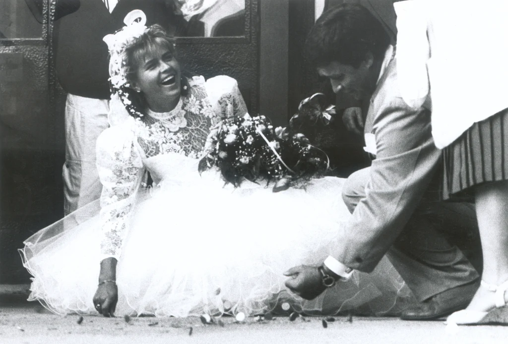 Ślub Doroty Tlałki i Christiana Mogore Panna młoda w sukni ślubnej uśmiecha się szeroko, klęcząc lub siedząc na ziemi. Obok niej mężczyzna w garniturze pomaga zebrać rozsypane monety, atmosfera jest radosna i lekko humorystyczna, a w tle widoczna jest część innych osób.