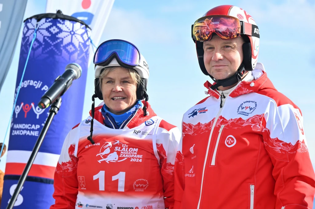 Małgorzata Tlałka-Długo z byłym prezydentem Andrzejem Dudą Dwie osoby w sportowych strojach narciarskich stoją obok siebie, uśmiechnięte, z numerem startowym oraz narciarskimi kaskami i goglami, w tle widoczny mikrofon i baner wydarzenia sportowego.