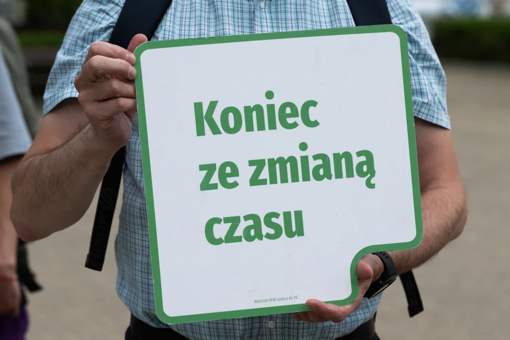 Kiedy będzie ostatnia zmiana czasu? Obowiązujące regulacje wywołują sprzeciw mieszkańców UE. Osoba w koszuli w kratę trzyma tabliczkę z napisem 'Koniec ze zmianą czasu' na jasnym tle, wokół widoczna jest zielona ramka.