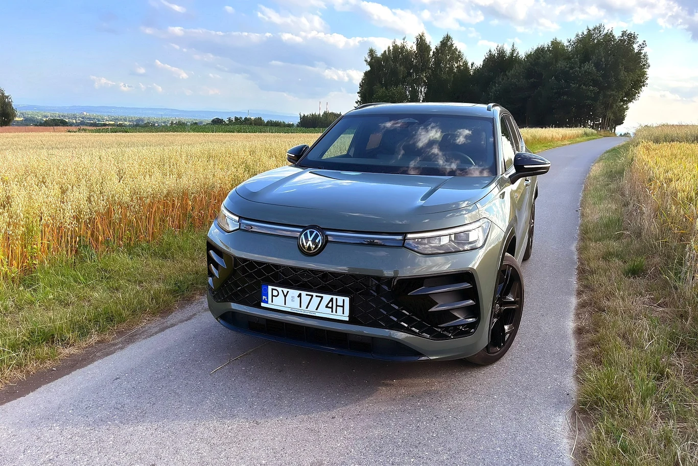 Volkswagen Tayron z przodu do złudzenia przypomina Tiguana / Fot: Tomasz Szpil Nowoczesny szary SUV marki Volkswagen stoi na wąskiej asfaltowej drodze otoczonej polami zboża, w tle widoczne są drzewa i otwarty krajobraz.