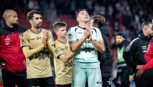 Grupa piłkarzy ubranych w stroje sportowe stoi na murawie stadionu, zawodnicy klaszczą i wyrażają emocje po zakończonym meczu, w tle widoczne są inne osoby i trybuny stadionu.