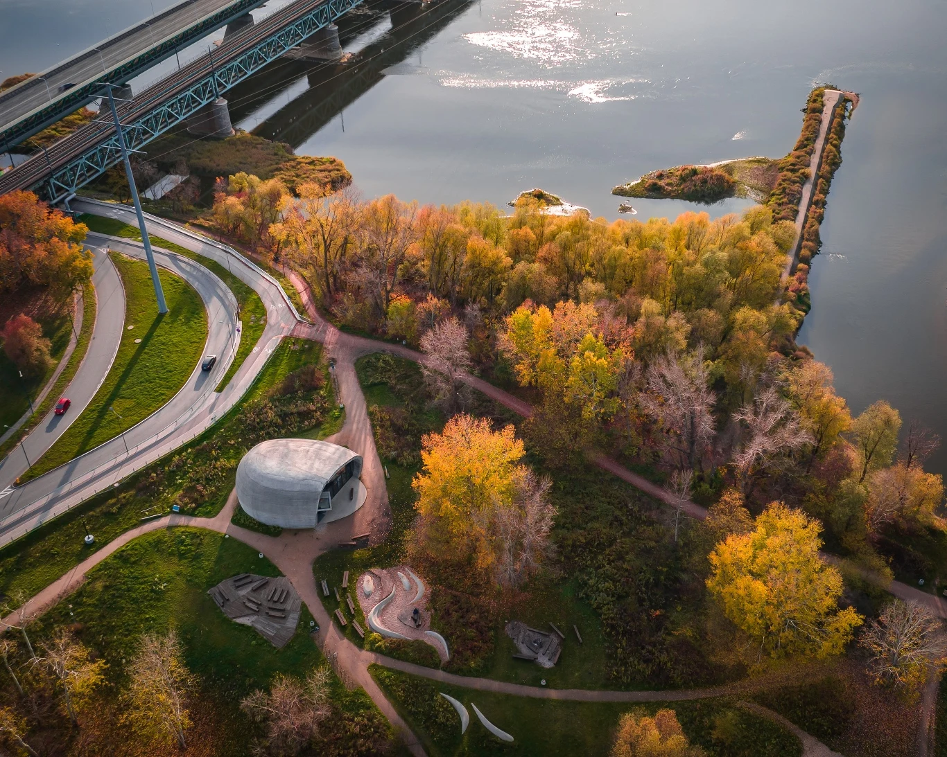 Pawilon Edukacyjny "Kamień" w Warszawie Park z kolorowymi drzewami jesienią, nowoczesny pawilon i ścieżki spacerowe prowadzące do rzeki, w tle most kolejowy i fragment wału oddzielający wodę.