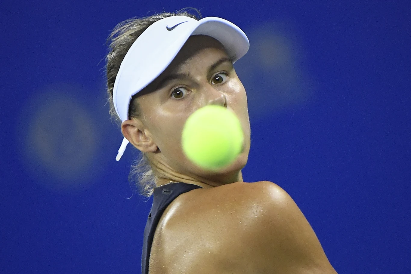 Kobieta w sportowym stroju i białym daszku śledzi z koncentracją zbliżającą się piłkę tenisową na tle niebieskiego kortu.