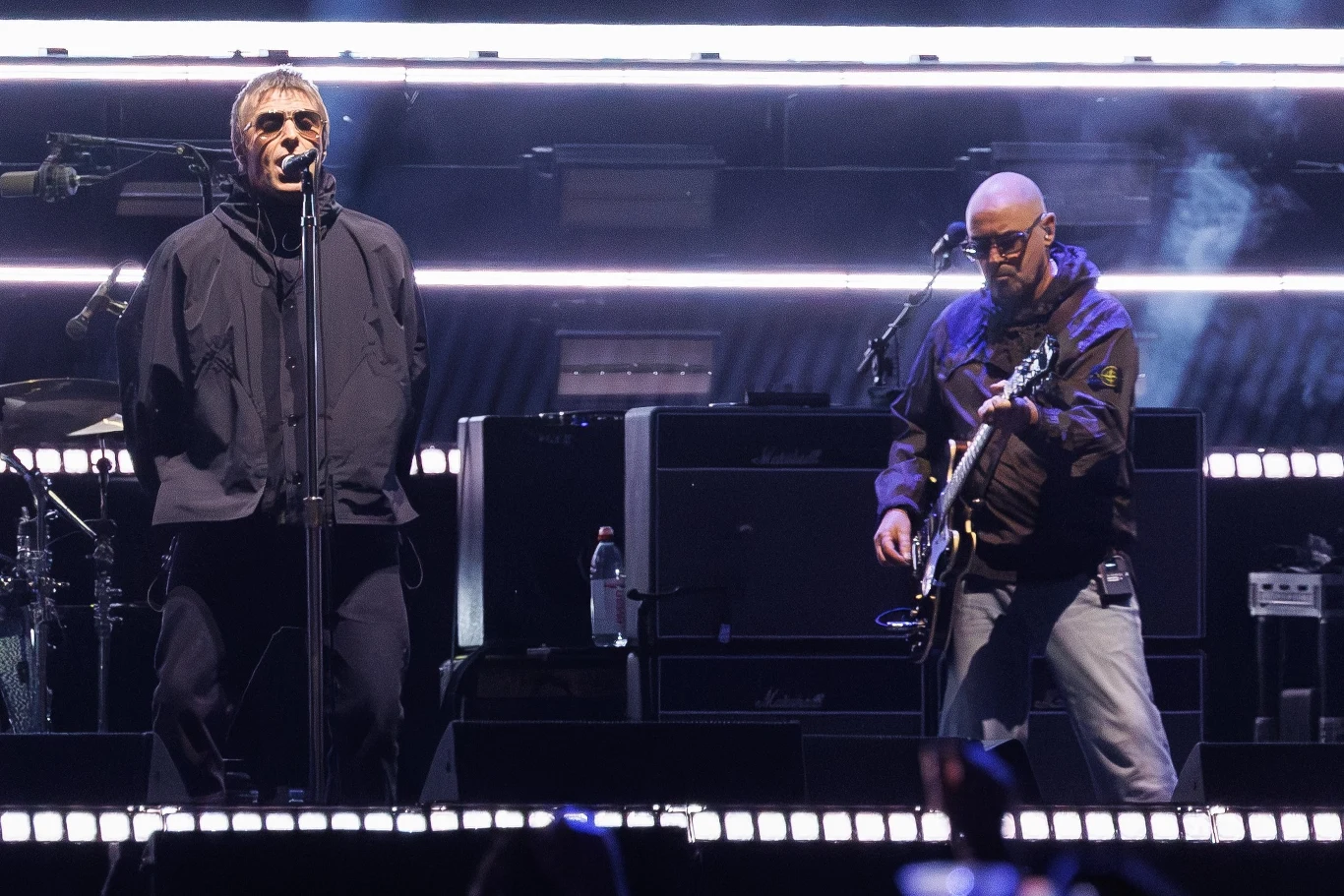Liam Gallagher i Paul "Bonehead" Arthurs z Oasis na scenie Dwóch muzyków na scenie, wokalista stoi przy mikrofonie w ciemnej kurtce, obok gitarzysta w okularach gra na gitarze elektrycznej, w tle widoczne wzmacniacze i efektowne oświetlenie sceniczne.
