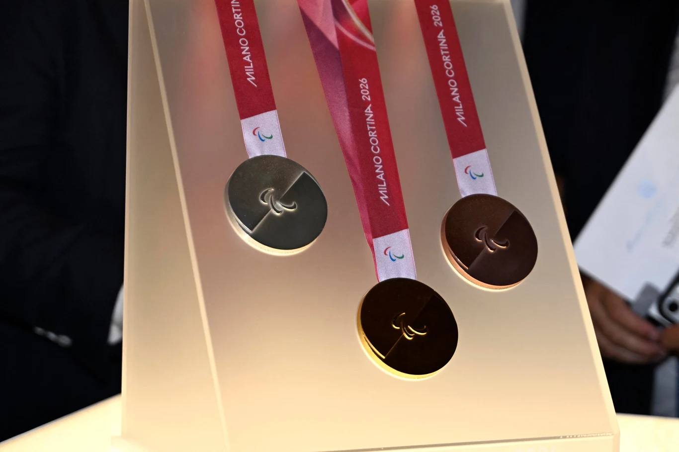 Tak wyglądają medale zimowych igrzysk olimpijskich 2026 Trzy medale olimpijskie z różnych kruszców zawieszone na czerwonych wstążkach z napisem Milano Cortina 2026 umieszczone na jasnej ekspozycji.