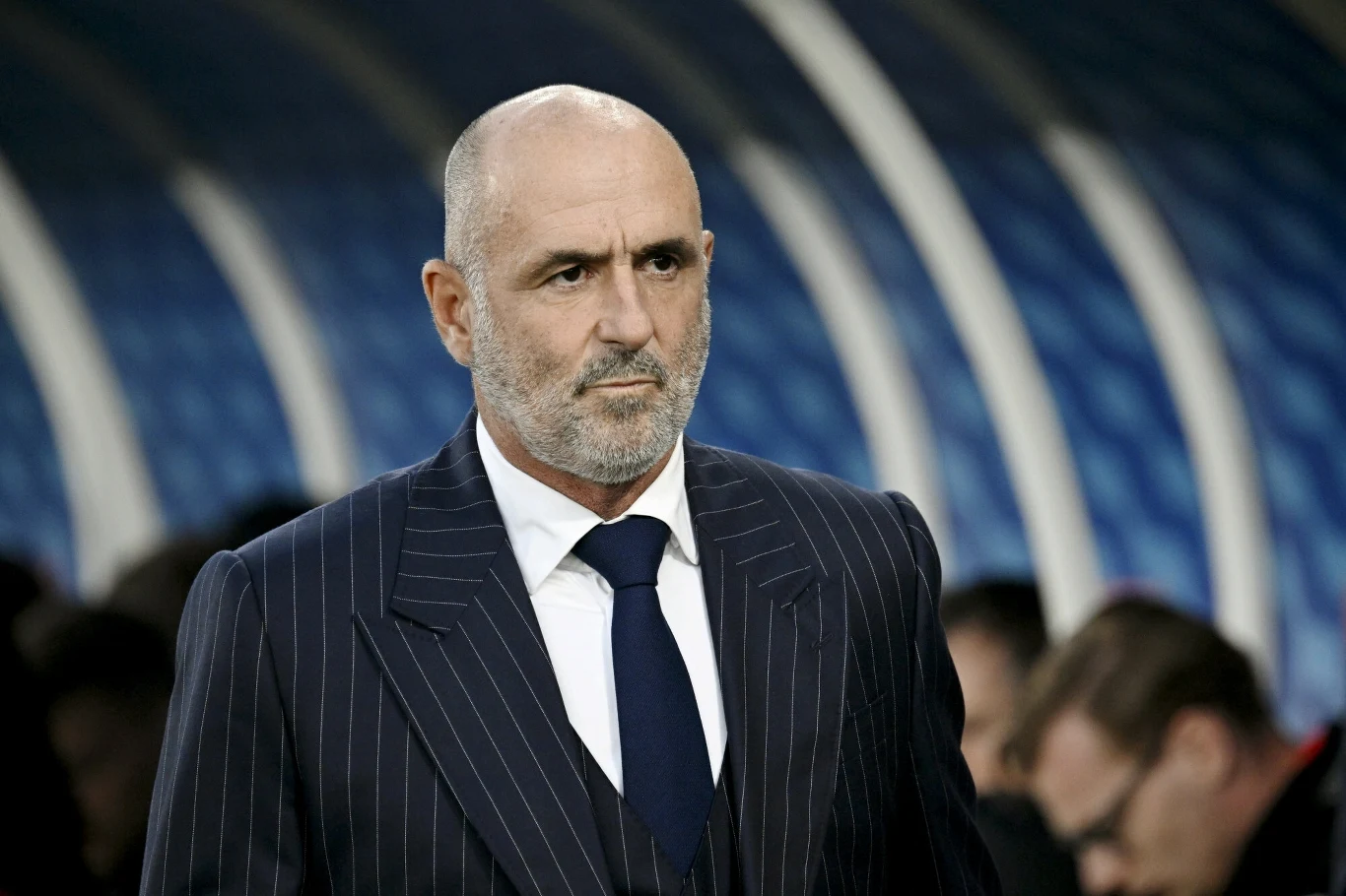 Mężczyzna w średnim wieku o siwych włosach i brodzie, ubrany w elegancki, pasiasty garnitur z krawatem, stoi na tle stadionu, sprawiając wrażenie skupionego i poważnego.