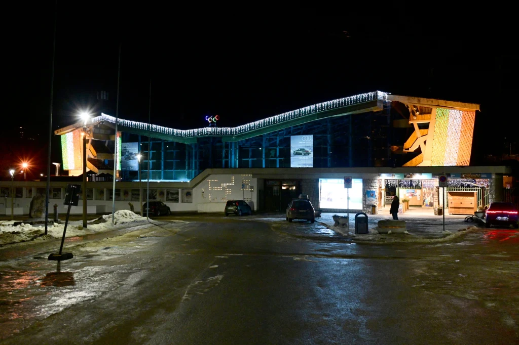 Cortina Curling Olympic Stadium - arena zmagań curlerów na ZIO 2026 Oświetlony skocznia narciarska nocą, na parkingu przed obiektem znajdują się zaparkowane samochody i niewielka ilość resztek śniegu, budynek wyróżnia się nowoczesną architekturą i jasnym podświetleniem