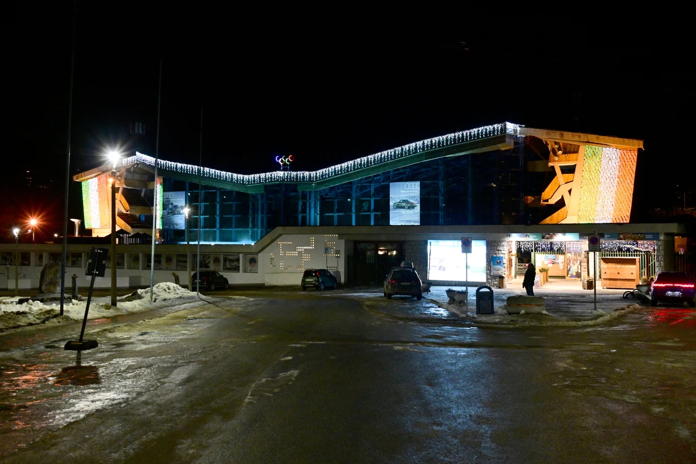Cortina Curling Olympic Stadium - arena zmagań curlerów na ZIO 2026 Oświetlony skocznia narciarska nocą, na parkingu przed obiektem znajdują się zaparkowane samochody i niewielka ilość resztek śniegu, budynek wyróżnia się nowoczesną architekturą i jasnym podświetleniem