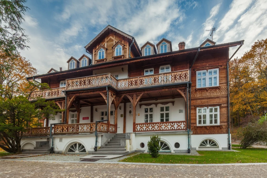 Czujecie alpejski klimat? Duży, zabytkowy drewniany budynek z ozdobnymi balkonami, białą podmurówką i wielospadowym dachem, otoczony jesiennym krajobrazem i drzewami.