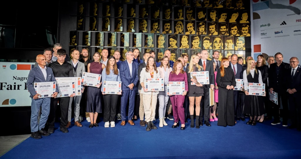 Grupa elegancko ubranych osób stojących razem na niebieskim dywanie, większość z nich trzyma dyplomy lub tablice z napisem 'Dyplom', na ścianie w tle portrety sportowców oraz logotypy partnerów wydarzenia.