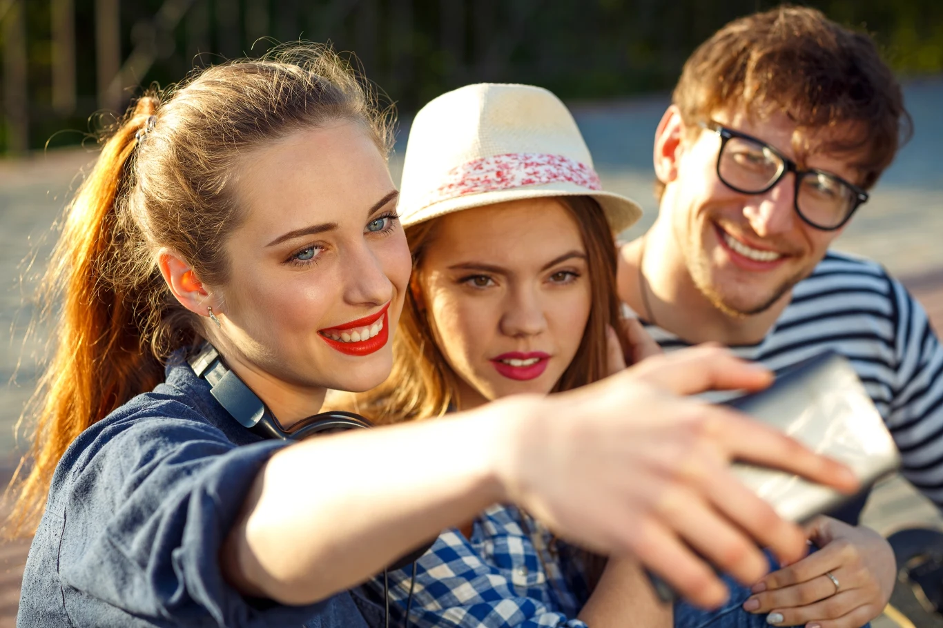 Trzy młode osoby uśmiechają się i robią wspólne selfie na zewnątrz w słoneczny dzień, na pierwszym planie wyraźne są uśmiechy, świeże kolory i luźna atmosfera.