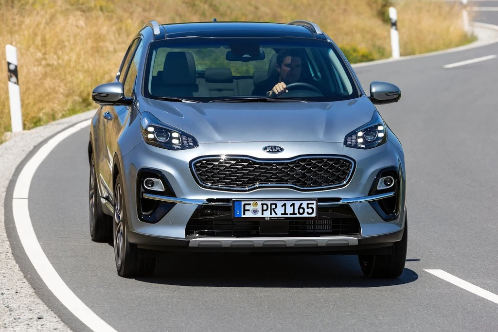 Srebrny samochód Kia Sportage poruszający się po asfaltowej, krętej drodze na tle suchej trawy, widoczny z przodu z wyraźnym grillem i światłami, za kierownicą siedzi jedna osoba.