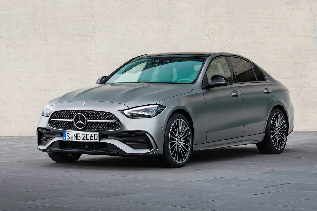 Srebrny samochód osobowy marki Mercedes-Benz klasy C na tle jasnej betonowej ściany, z wyraźnie widocznym przednim grillem, nowoczesnymi reflektorami i charakterystycznymi felgami.