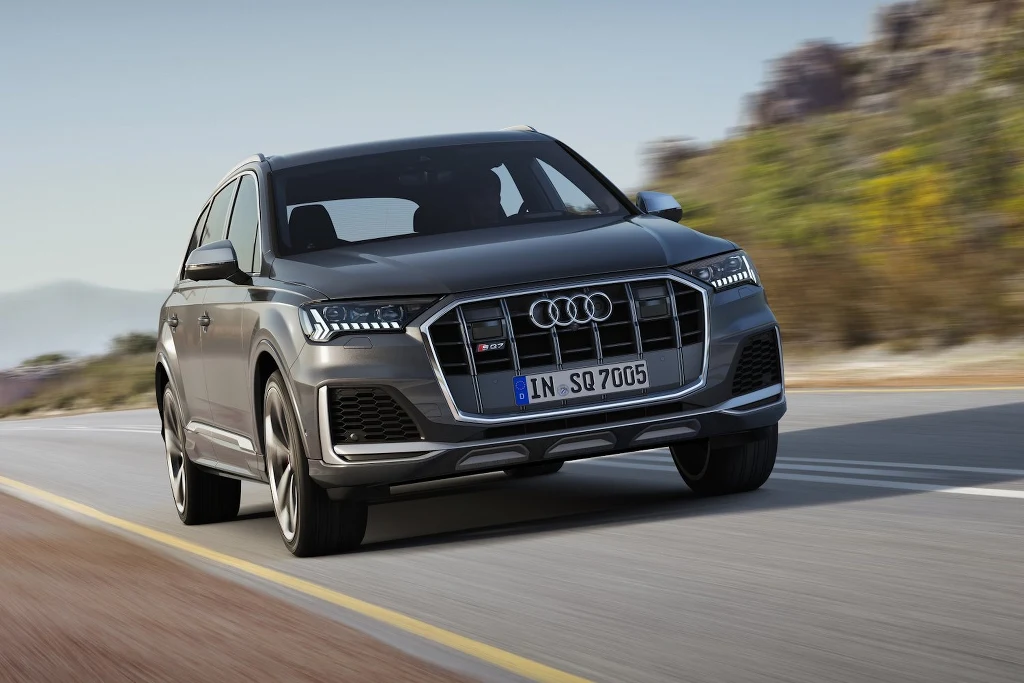 Szary SUV marki Audi jedzie po asfaltowej drodze, widoczny z przodu pod lekkim kątem, w tle rozmyty krajobraz z skałami i roślinnością.