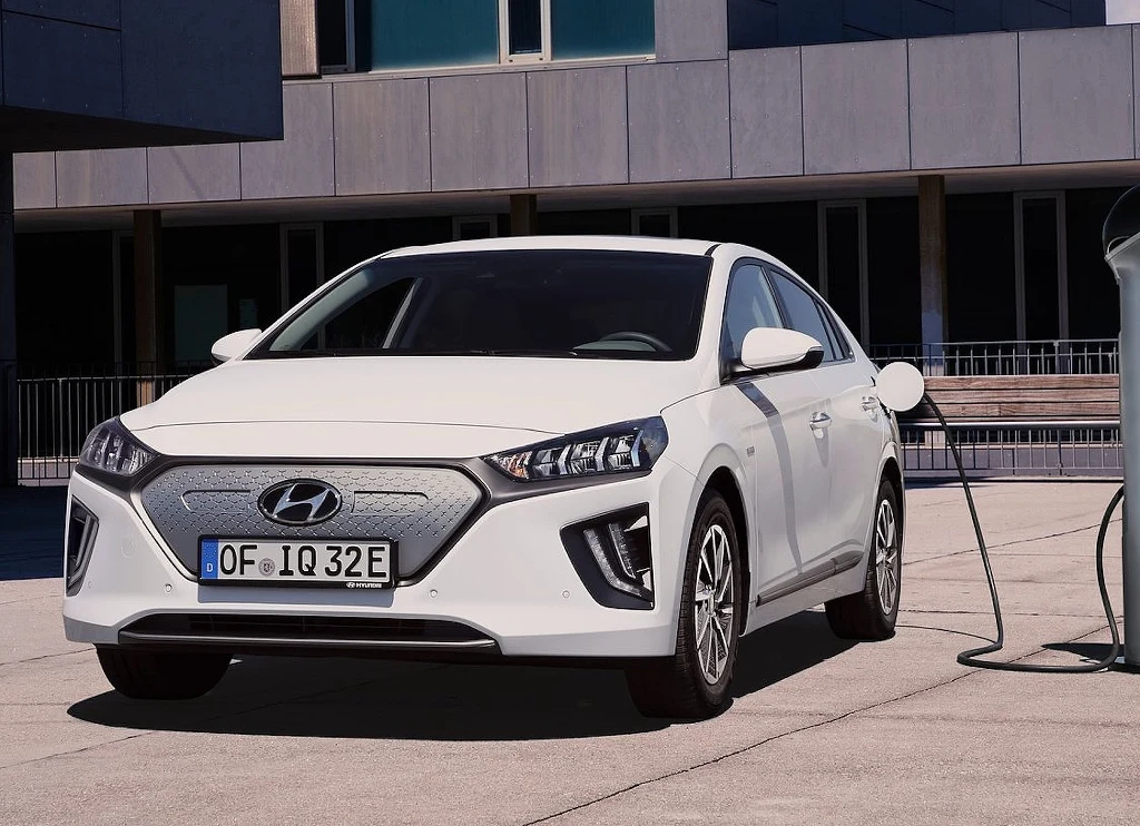 Biały samochód marki Hyundai Ioniq stoi zaparkowany przy stacji ładowania pojazdów elektrycznych na tle nowoczesnego budynku z dużymi oknami.