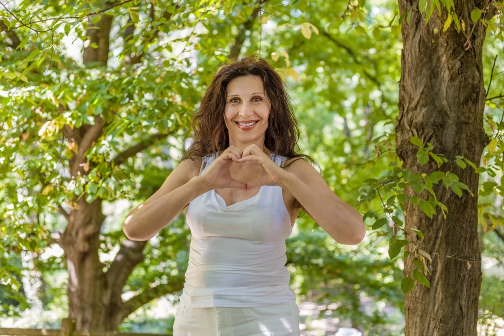 Jak działa post przerywany? Kobieta w białej bluzce stoi w parku wśród drzew, uśmiecha się i pokazuje dłońmi gest serca.