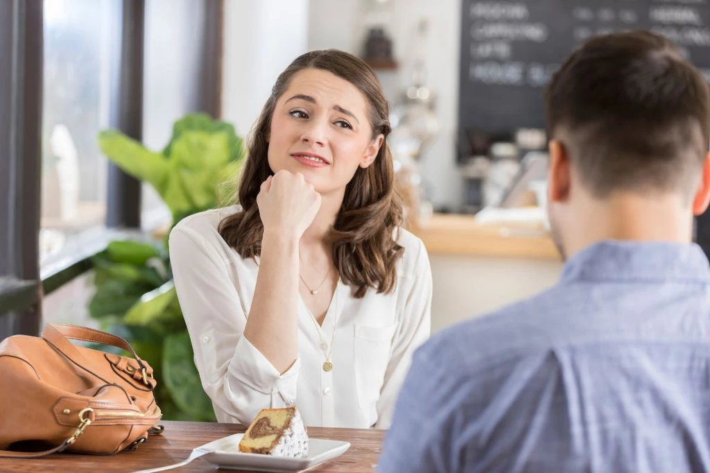 Kobieta w białej koszuli siedzi przy stoliku w kawiarni, patrząc z wyraźnym zniecierpliwieniem lub niezadowoleniem na siedzącego naprzeciw niej mężczyznę; na stole kawałek ciasta i torebka.