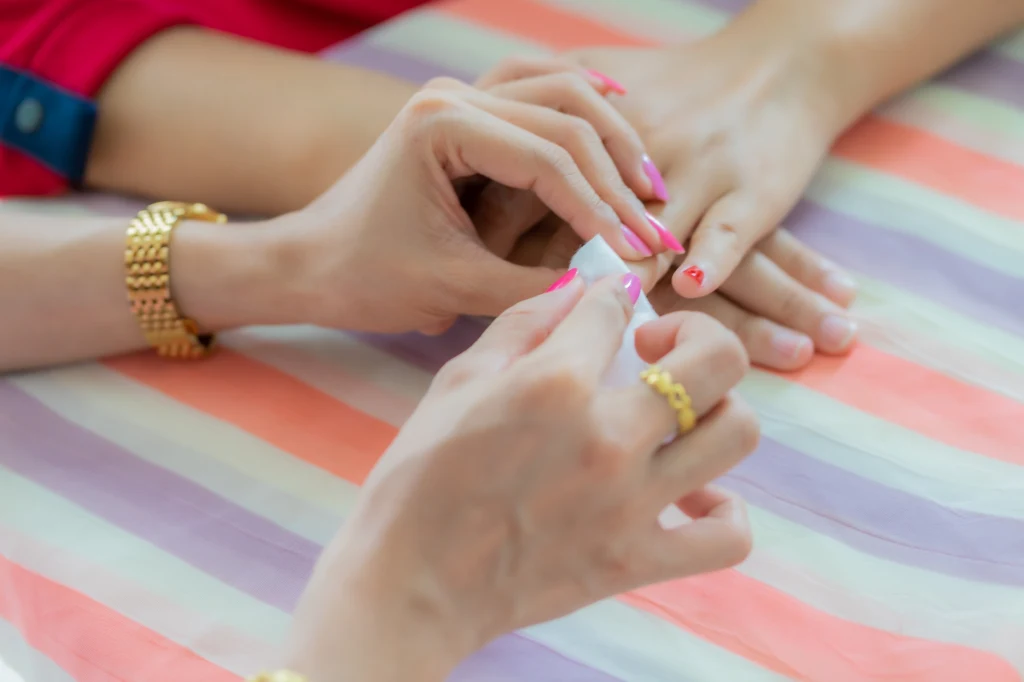 Jak działa manicure japoński i dla kogo jest najlepszym wyborem? Ręce dwóch osób podczas wykonywania manicure, jedna z nich maluje paznokcie drugiej lakierem w jasnym kolorze, dłonie leżą na kolorowym, paskowanym materiale.