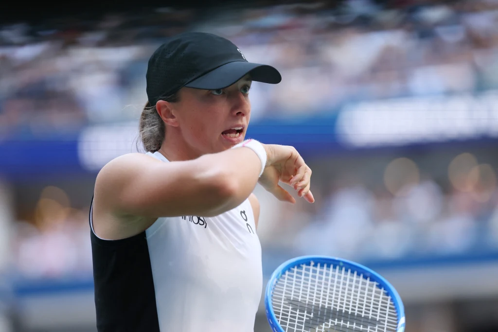 Iga Świątek Tenisistka ubrana w sportowy strój i czapkę z daszkiem trzyma niebieską rakietę, wykonując gest ręką, w tle rozmyte trybuny z kibicami.