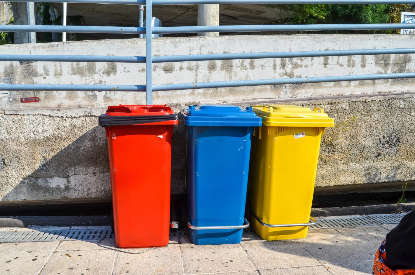 W Polsce czerwony pojemniki na śmieci możemy spotkać coraz częściej Trzy plastikowe pojemniki na odpady stoją obok siebie na chodniku, każdy w innym kolorze: czerwonym, niebieskim i żółtym, tło stanowi betonowy mur i metalowa barierka.
