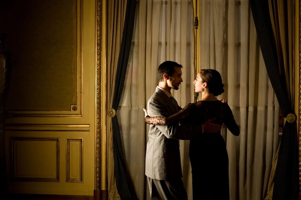 Elegancka para tańczy w gustownym, historycznym wnętrzu, światło subtelnie oświetla ich sylwetki na tle zasłoniętych, wysokich okien.