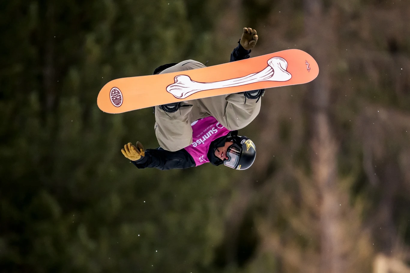 Mistrzem olimpijskim w konkurencji Big Air w 2022 roku został Yiming Su Sportowiec w kasku i goglach wykonujący efektowną akrobację na snowboardzie w powietrzu, w tle rozmyty las, a na desce wyraźny, duży rysunek kości.