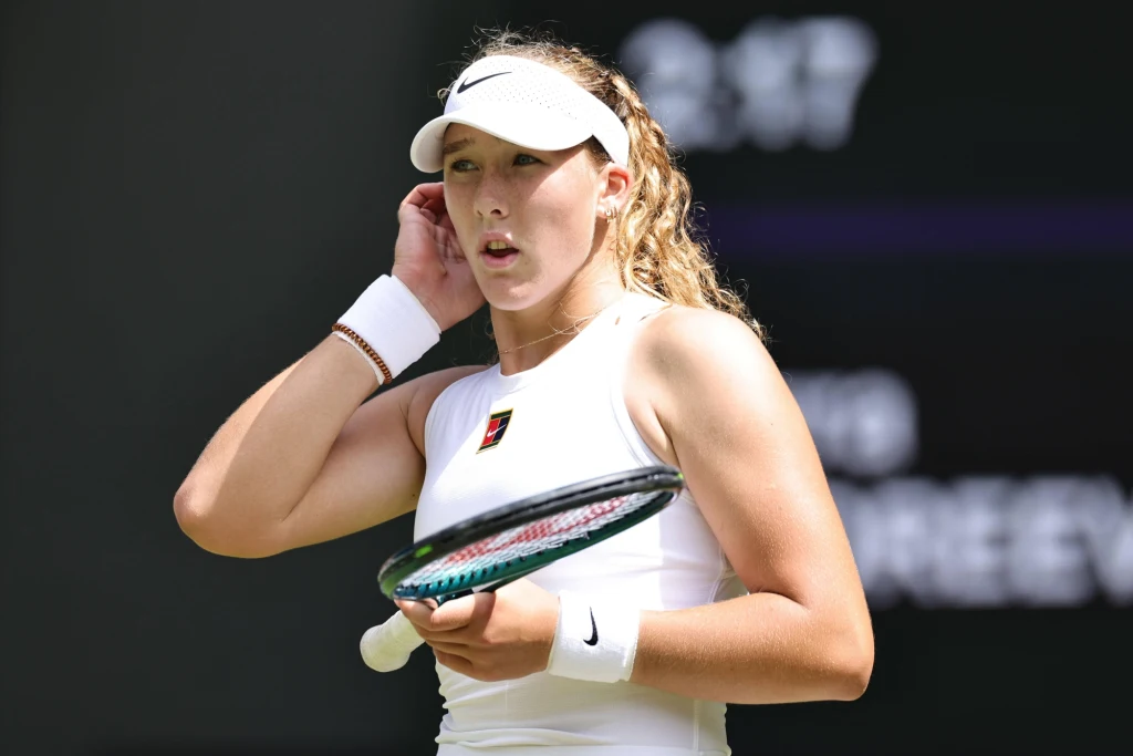 Mirra Andriejewa Tenisistka w białym stroju sportowym i daszku trzyma rakietę na tle rozmazanej tablicy wyników, koncentrując się przed kolejną akcją.