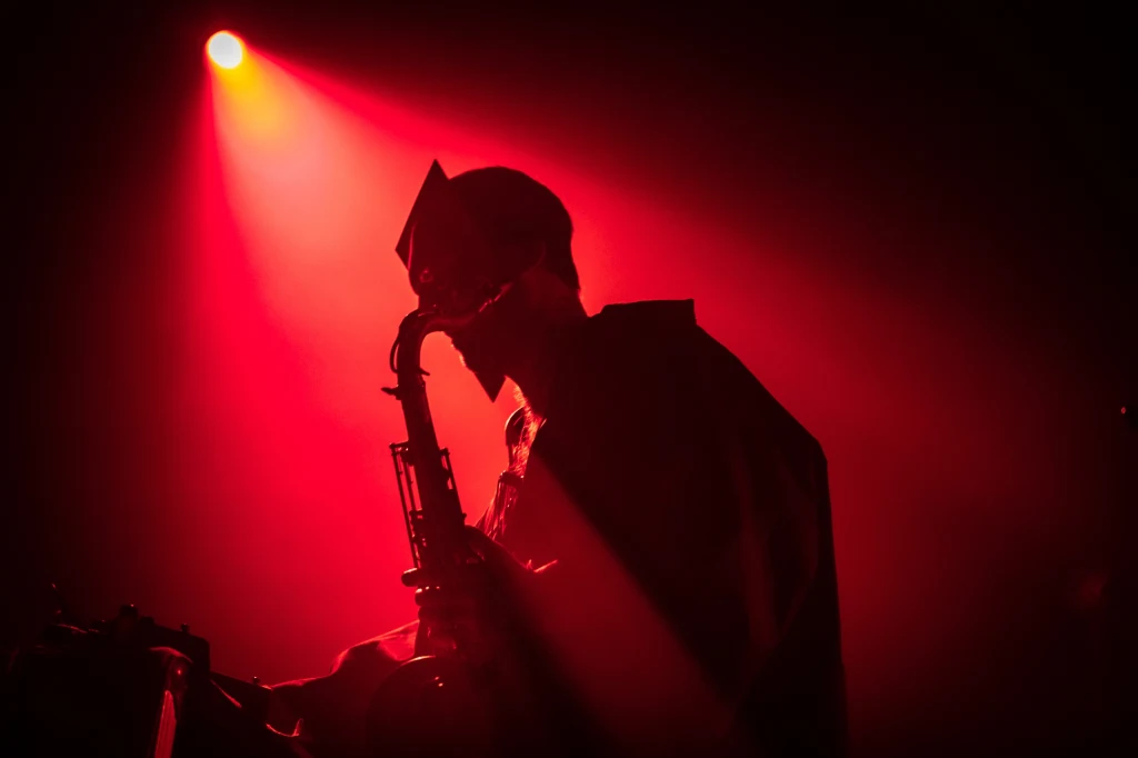 Monsters of Jazz: Nene Heroine Sylwetka muzyka grającego na saksofonie w czerwonym świetle sceny, atmosfera tajemnicza, podkreślona intensywnym, rozproszonym oświetleniem.