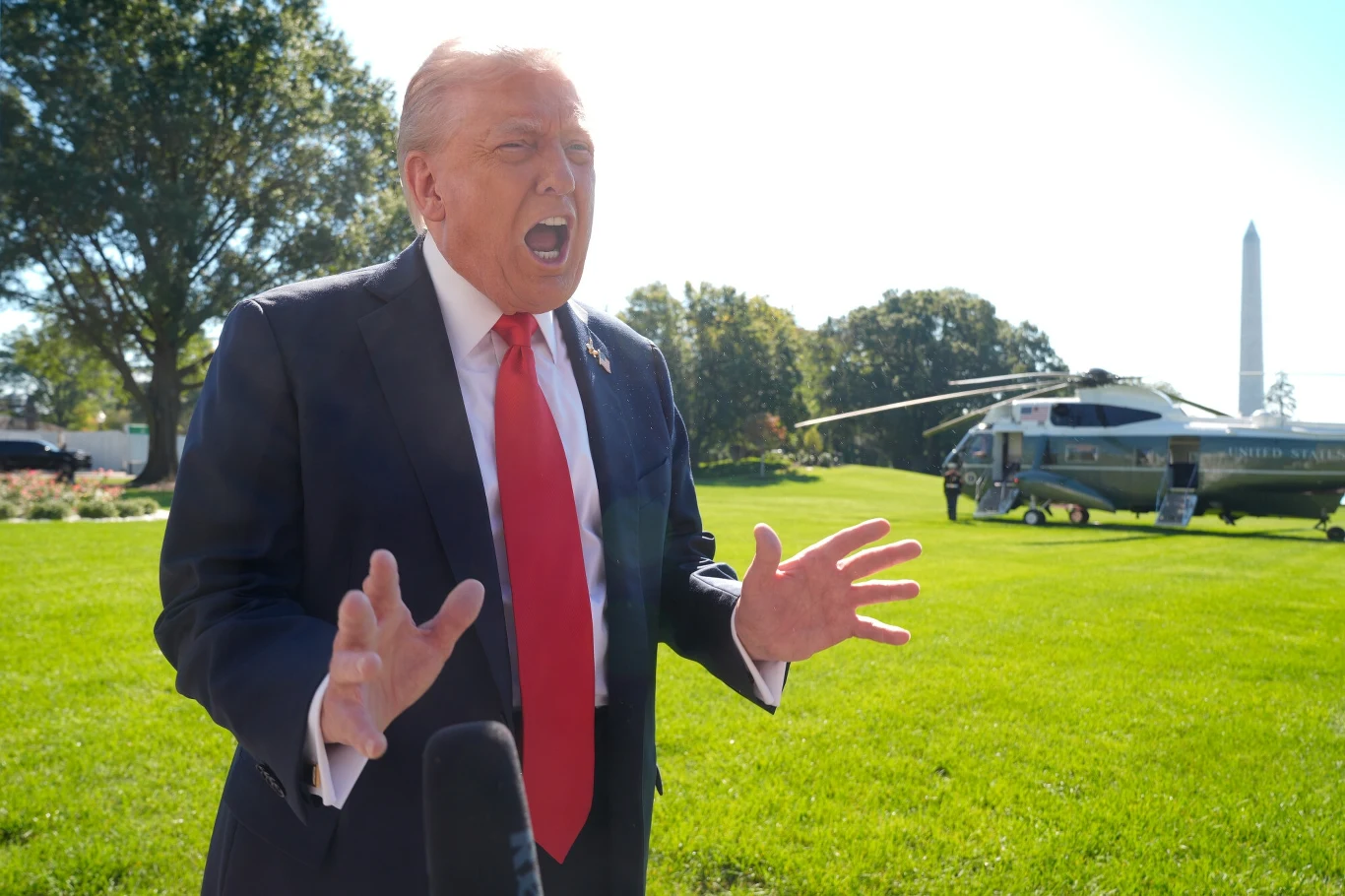 Mężczyzna w garniturze i czerwonym krawacie gestykuluje podczas przemowy na zielonym trawniku, w tle widać helikopter i monument.