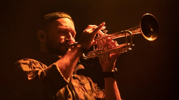Mężczyzna w wojskowej koszuli i czapce grający na trąbce, oświetlony ciepłym, pomarańczowym światłem, z intensywnym wyrazem twarzy skupionym na grze.