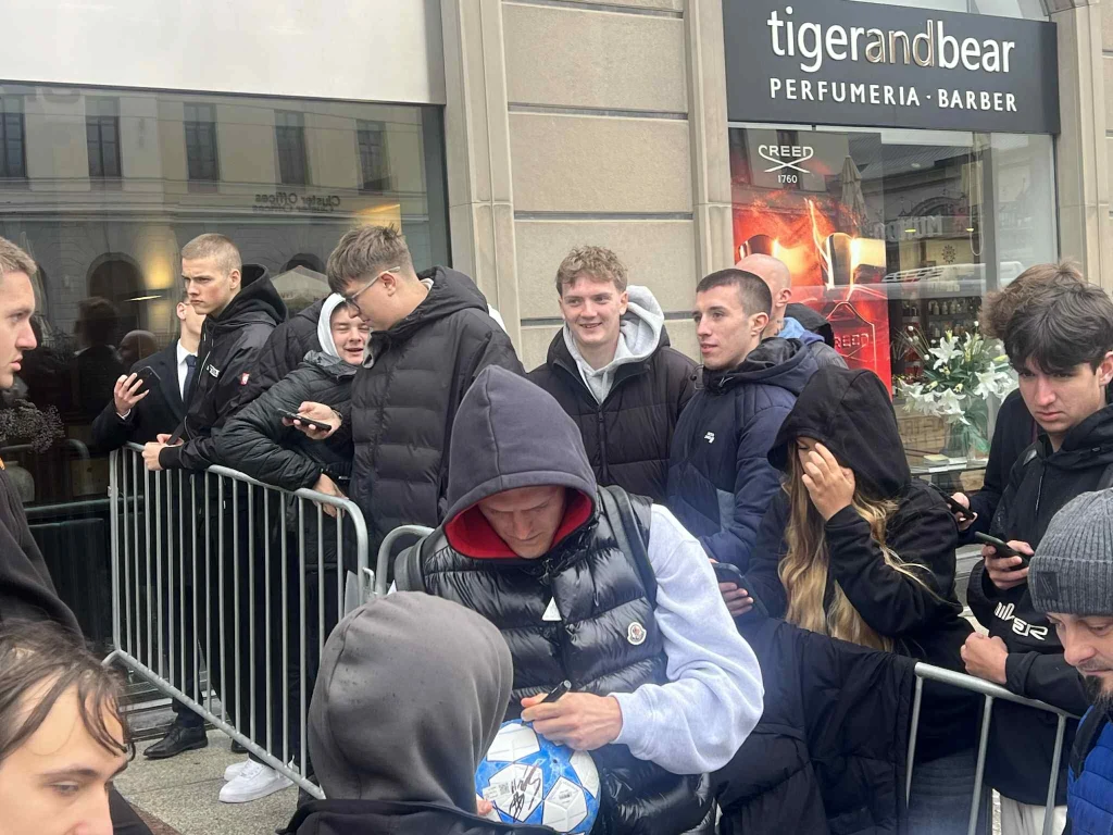 Karol Świderski rozdaje autografy Grupa młodych osób w zimowych kurtkach stoi oraz siedzi przy metalowych barierkach przed wejściem do perfumerii i barbershopu tigerandbear, niektórzy korzystają z telefonów, jedna osoba trzyma piłkę.