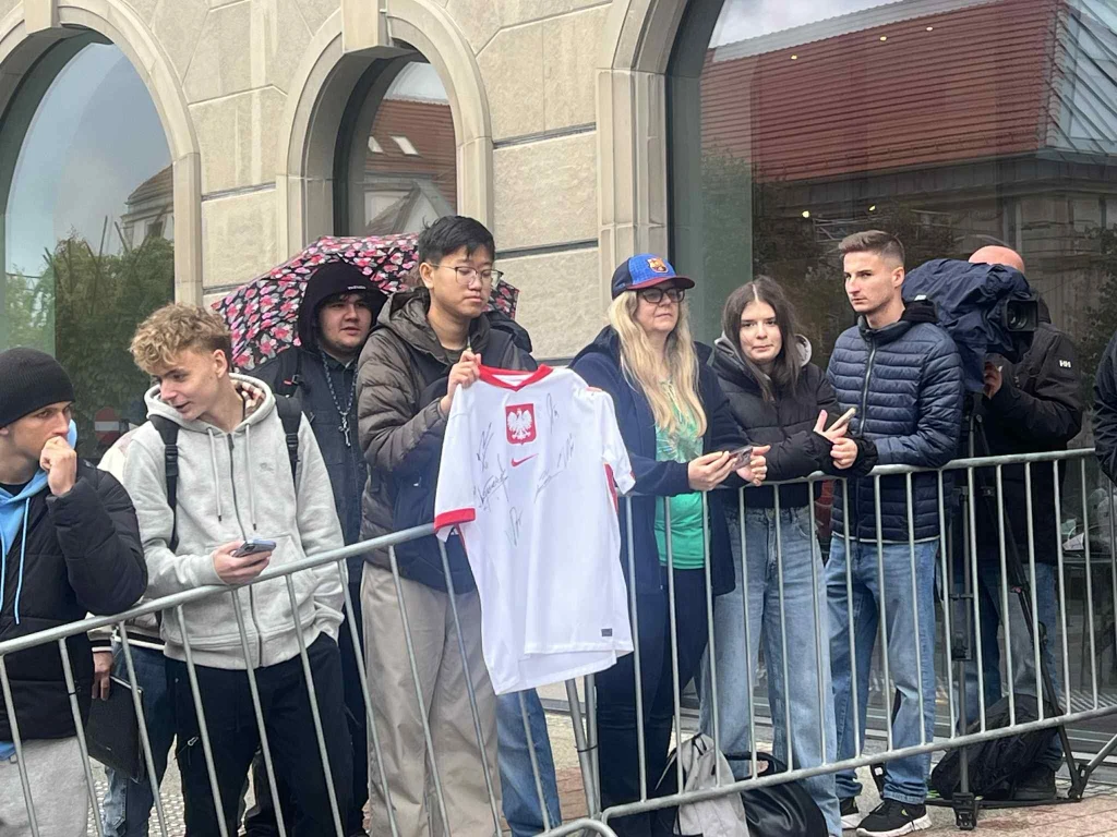 Kibice pod hotelem reprezentacji Polski Grupa młodych ludzi stoi za metalową barierką, jeden z nich trzyma koszulkę reprezentacji Polski z podpisami. Reszta osób rozmawia ze sobą lub robi zdjęcia telefonem, niektórzy trzymają parasole. W tle widoczne są okna i fragment budynku.