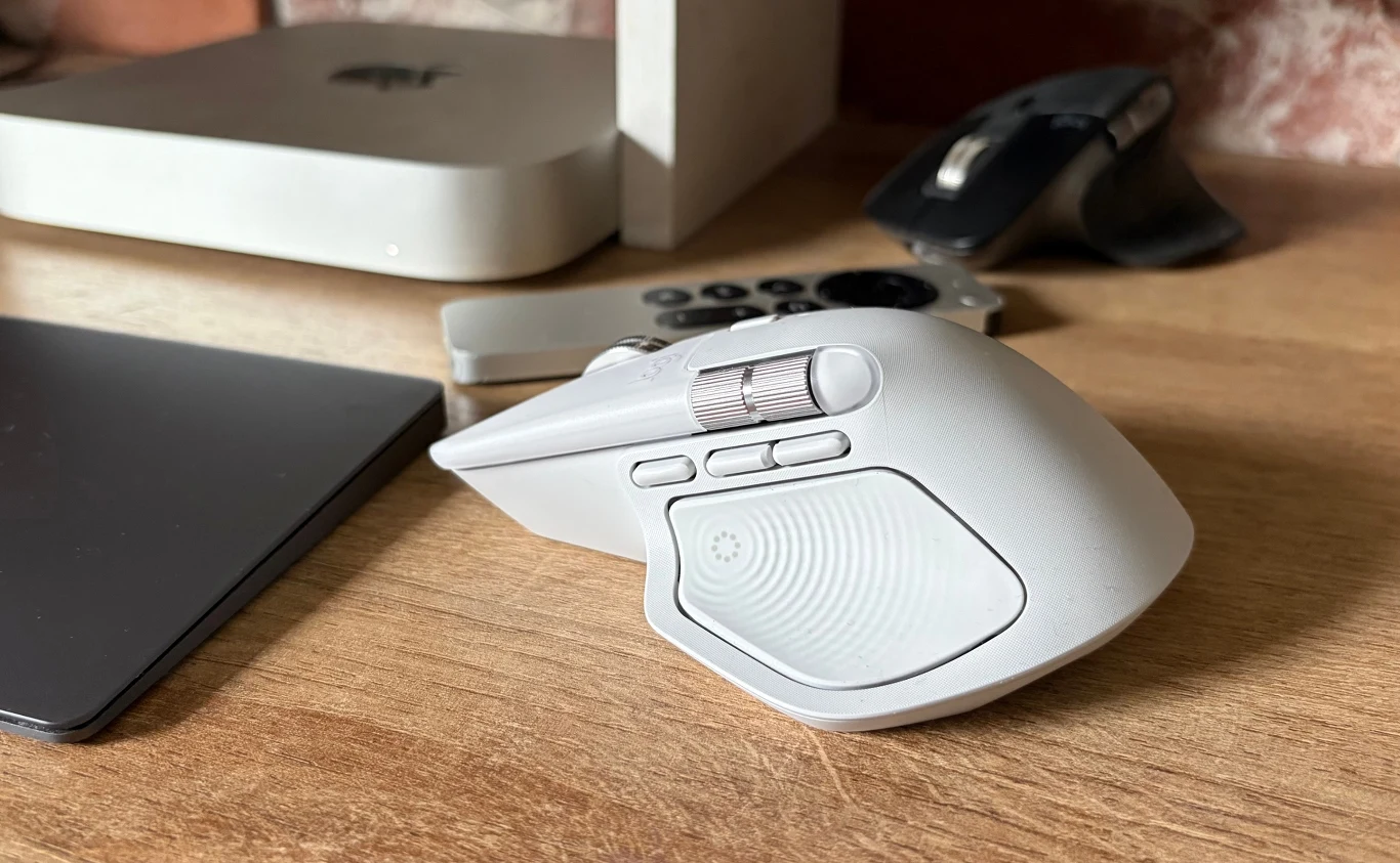 Test Logitech MX Master 4. Kultowa mysz komputerowa w nowej odsłonie. Biała, ergonomiczna mysz komputerowa z dodatkowymi przyciskami i metalową rolką, ustawiona na drewnianym biurku obok touchpada, pilota, czarnej myszy oraz części komputera stacjonarnego.