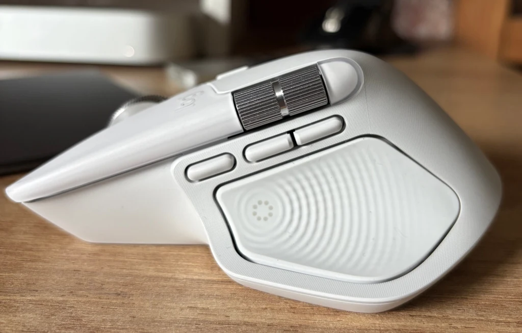 Logitech MX Master 4 ma nowy przycisk dla Actions Ring. Bezprzewodowa myszka komputerowa w jasnoszarym kolorze, z widocznym bocznym rolką oraz trzema przyciskami, leży na drewnianej powierzchni biurka.