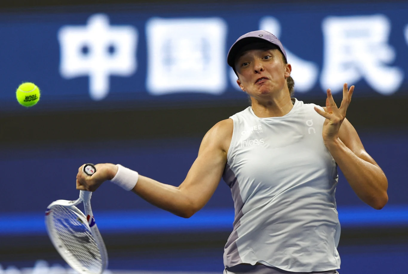 Iga Świątek turniej w Wuhan rozpoczyna od meczu z Marię Bouzkovą Tenisistka w białym stroju podczas dynamicznego uderzenia piłki rakietą podczas meczu, w tle nieostre napisy w języku chińskim.