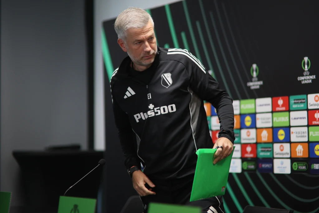 Trener w sportowym stroju z emblematami klubu piłkarskiego oraz logo sponsora stoi przy stole konferencyjnym, trzyma zieloną teczkę, w tle znajdują się ścianki z logotypami Ligi Konferencji i sponsorów.