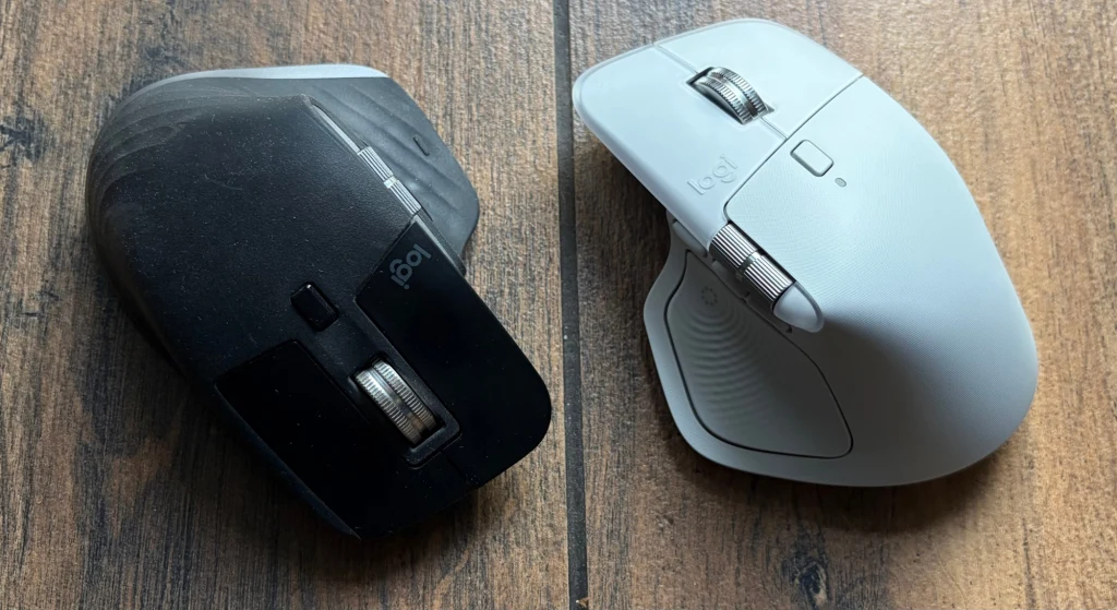Logitech MX Master 3 (po lewej) i MX Master 4 (po prawej). Dwie bezprzewodowe myszki komputerowe marki Logitech, jedna w kolorze czarnym, druga w białym, położone obok siebie na drewnianej powierzchni, podkreślając porównanie wyglądu i konstrukcji obu modeli.